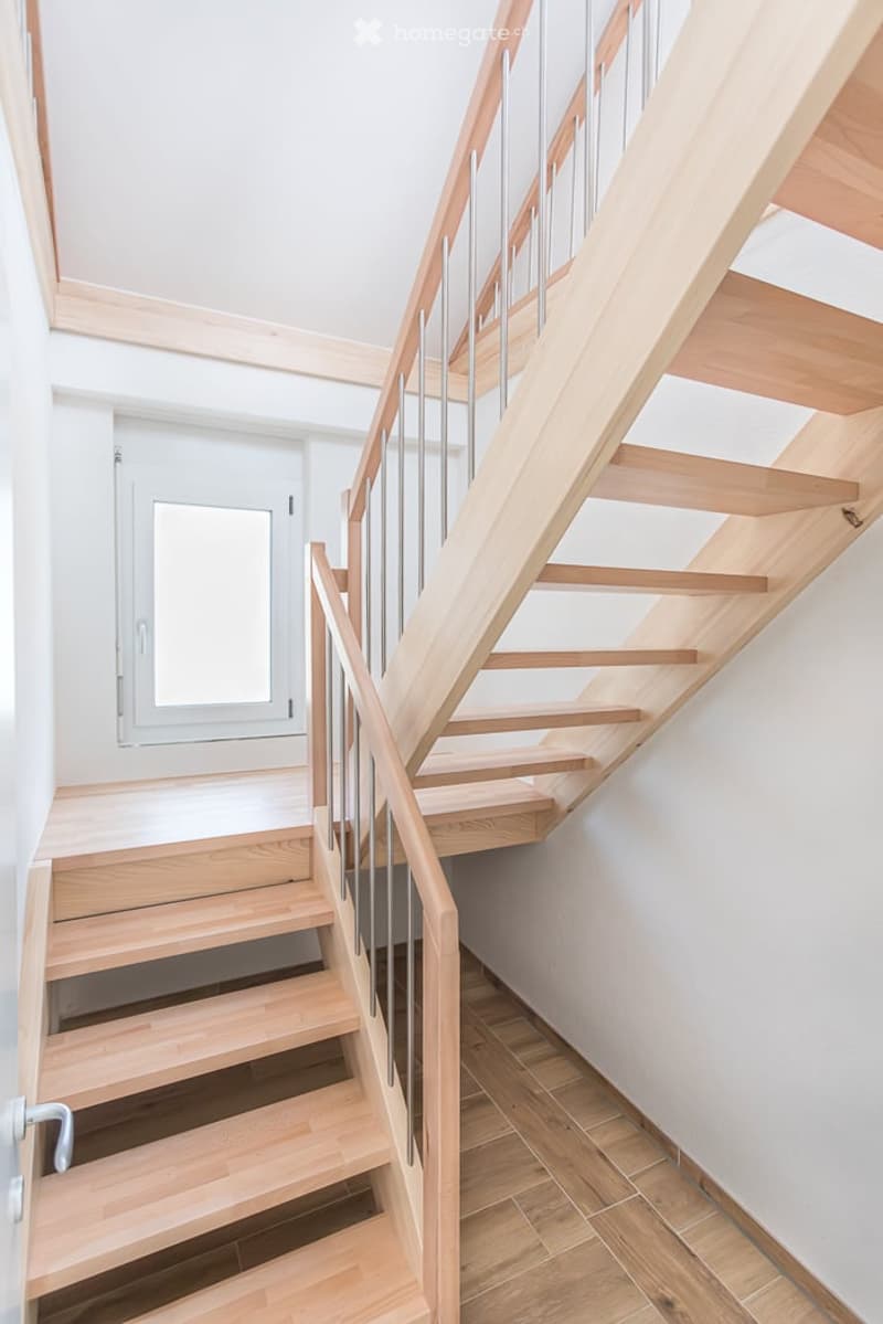 Modern wooden staircase with metal railing, light-colored wooden steps, white walls, and a window in the background.