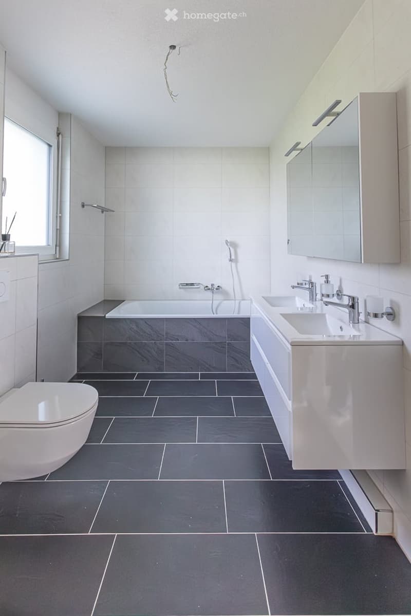 Modern bathroom with dark grey tiles, white fixtures, a bathtub, toilet, sink, and a mirrored cabinet.