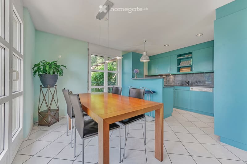 Bright dining area with a wooden table, six chairs, and a stool near the kitchen. The kitchen features turquoise cabinets, a dishwasher, and a countertop with a vase and plant. The room has large windows and modern lighting.
