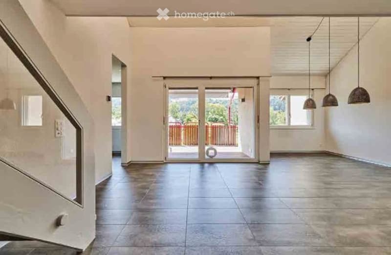 Empty hallway with tiled floor, white walls, staircase on the left, large glass doors leading to outdoor area, and hanging pendant lights.
