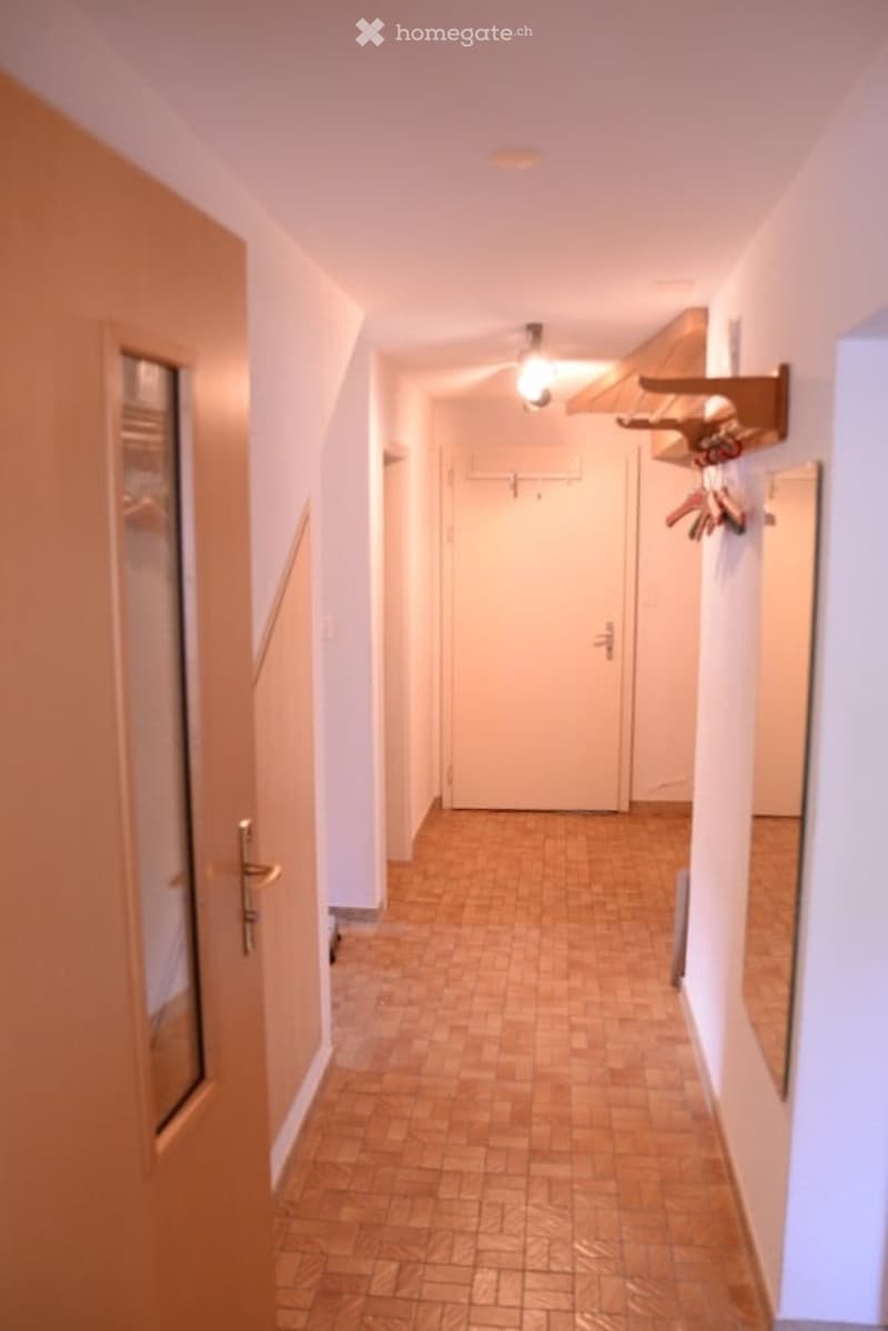 A narrow hallway with wooden tiled flooring, white walls, and a ceiling light fixture. A coat rack with hangers is mounted on the right wall, and a closed door is visible at the end of the hallway.