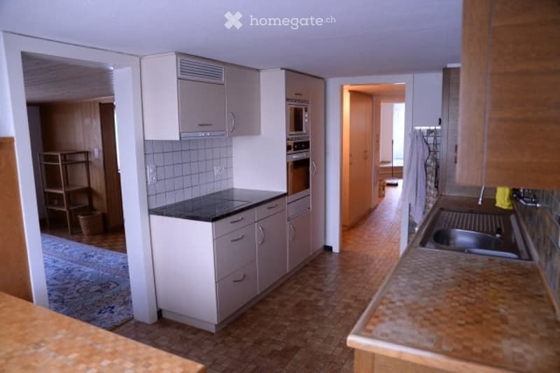 Modern kitchen with white cabinets, black countertop, stainless steel sink, and built-in oven. The floor is wooden, and there is a doorway leading to another room.