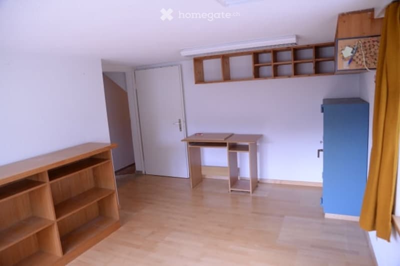 A small, unfurnished room with a wooden floor, white walls, and a single desk and shelf. The room is empty except for these items.