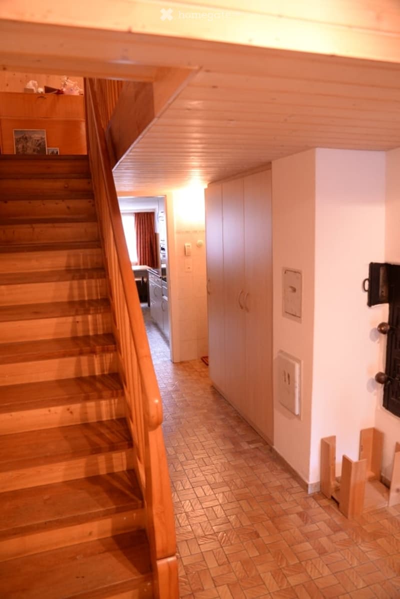 A well-lit hallway with wooden stairs, tiled floor, and built-in wooden cabinetry. The hallway leads to a room with a visible kitchen area in the background.