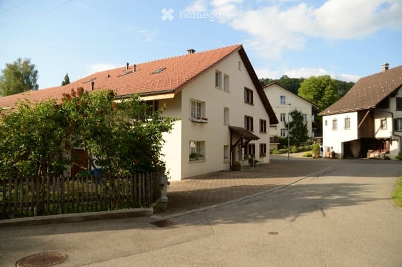 A two-story house with a red-tiled roof, white walls, and multiple windows, situated in a residential area with neighboring houses and greenery.