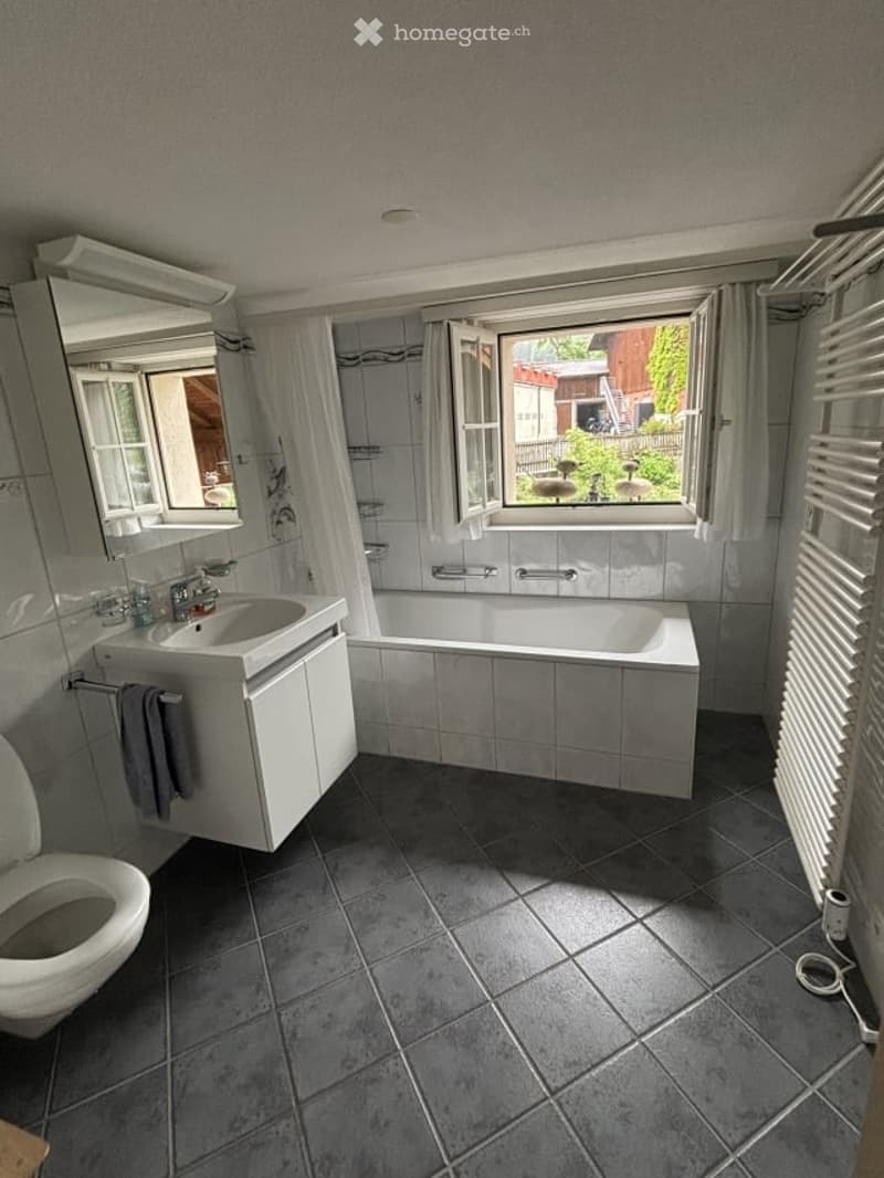 Bathroom with a white sink, bathtub, toilet, and mirror. The room features grey tiled walls and floor, with a window offering a view of the garden.