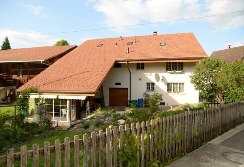 Two-story house with red-tiled roof, white exterior, and a small porch area with a table and chairs.