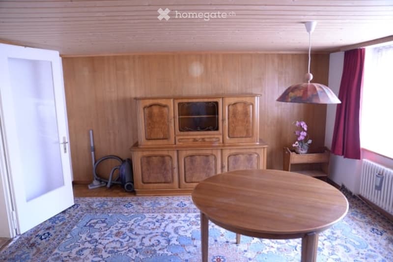 A wooden-paneled room with a large wooden cabinet, round wooden table, and a small side table with a flower vase. The room features a patterned carpet and a hanging lamp with a colorful shade.
