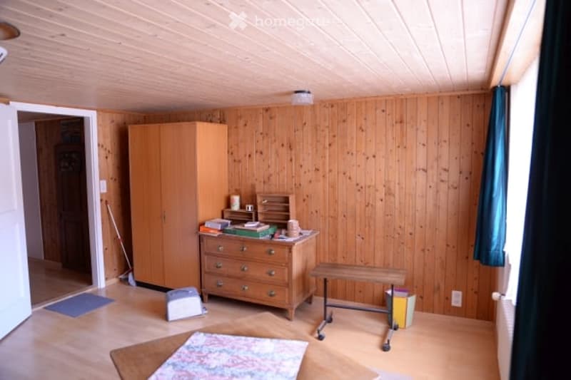 The room features wooden walls and flooring, with a wardrobe, chest of drawers, bench, table, and stool. There is also a small rug on the floor and a broom leaning against the wall.