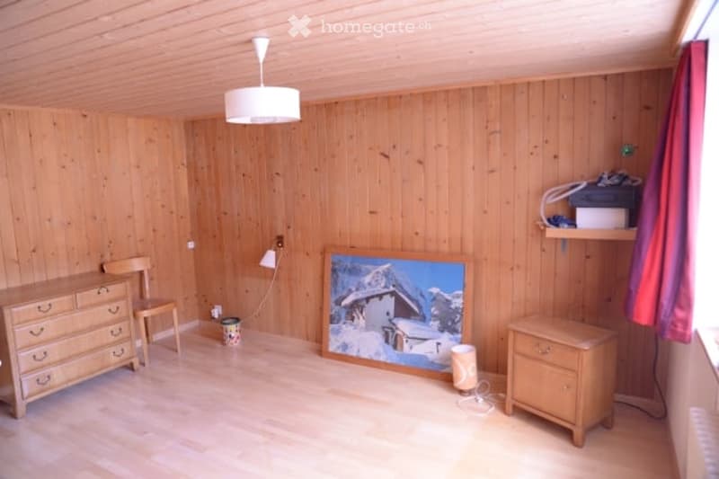 Cozy wooden-paneled room with a dresser, chair, picture frame, and a small stool. The room features a hanging lamp and a small trash bin.