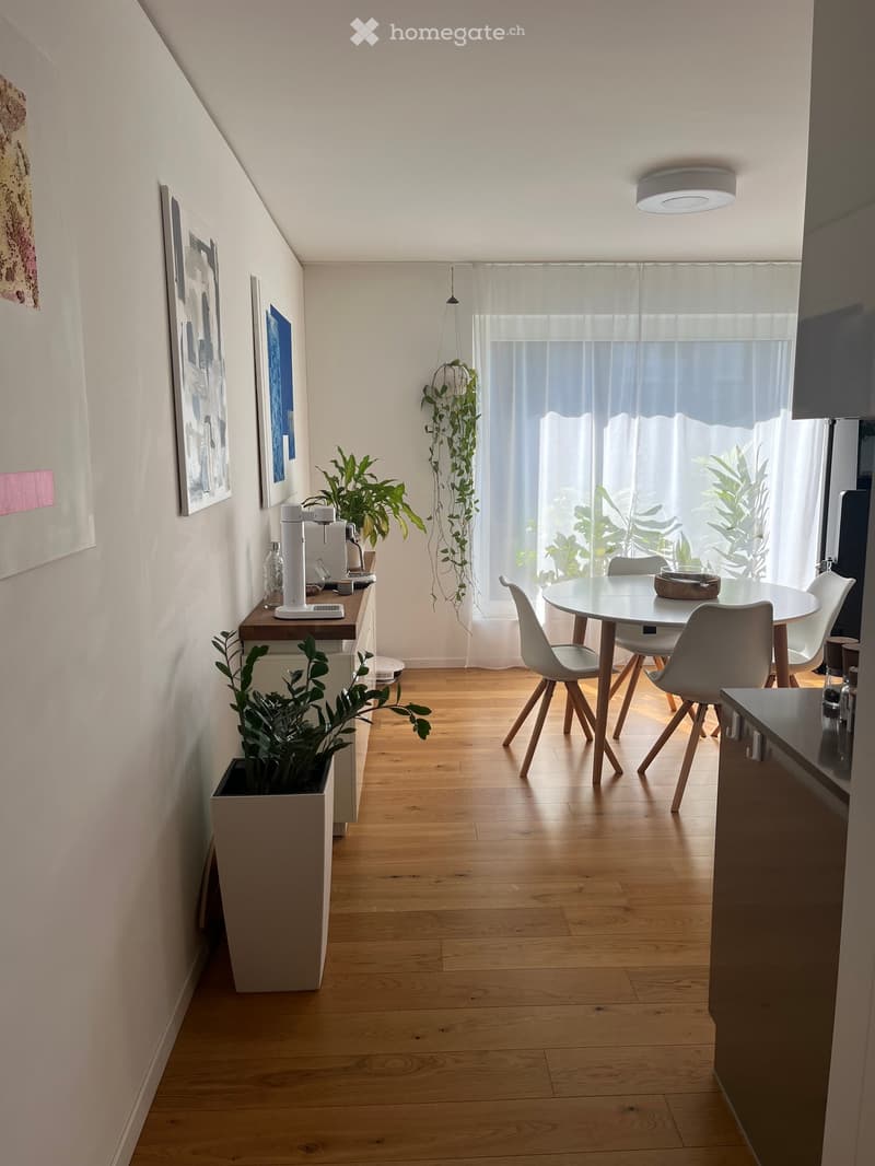 A modern, well-lit room with wooden flooring, a round dining table with four chairs, a coffee machine on a small counter, and several potted plants. The room features large windows with sheer curtains and artwork on the walls.