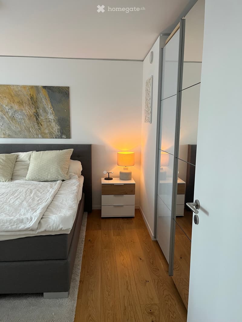 Modern bedroom with a wooden floor, a large bed with white bedding, a nightstand with a lamp, a mirrored wardrobe, and a painting on the wall.