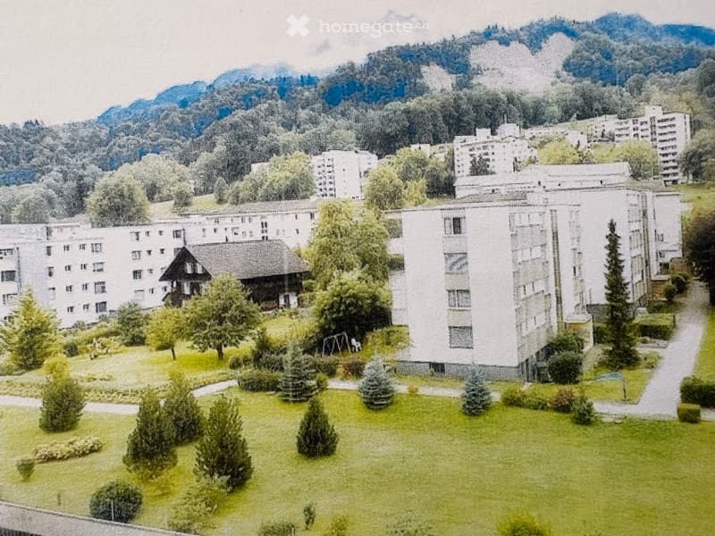 A large residential complex with multiple white apartment buildings, surrounded by green lawns, trees, and a mountainous backdrop.