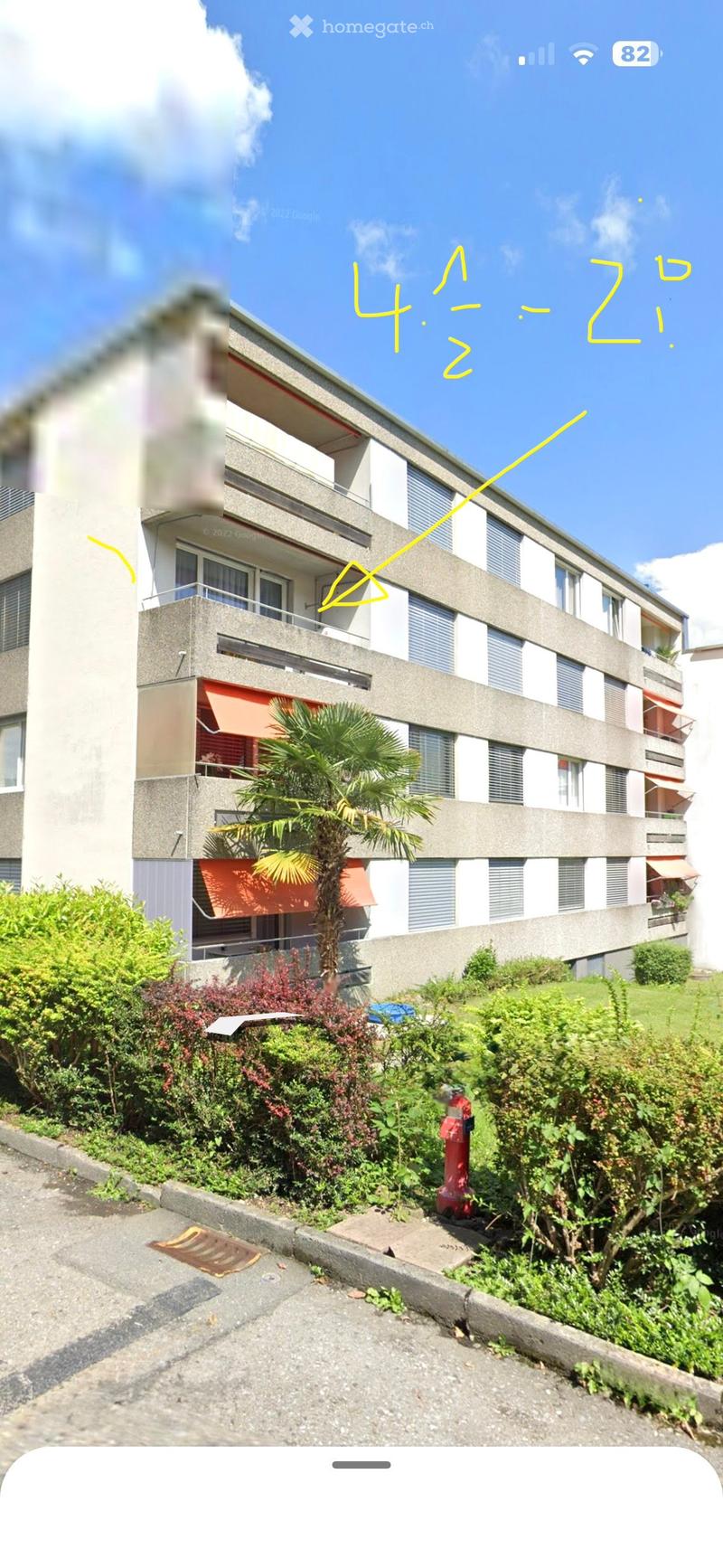 A multi-story residential building with balconies, surrounded by greenery and a palm tree. The building has a modern design with grey and white exterior walls.