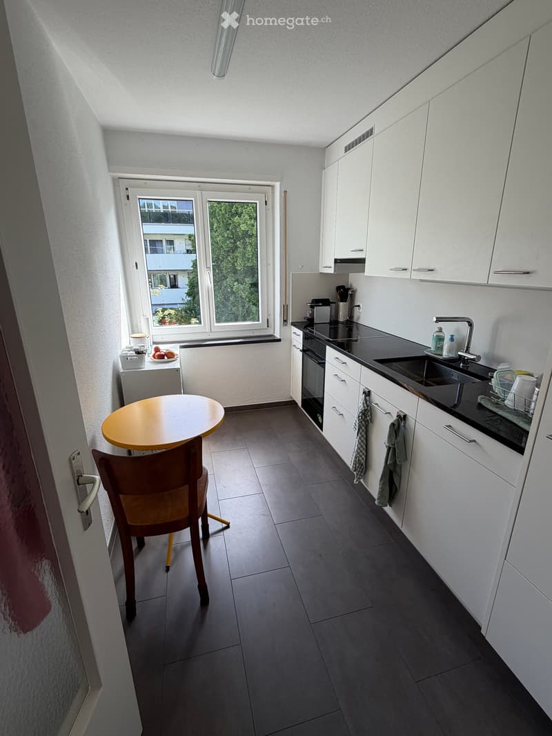 Modern kitchen with white cabinets, dark countertops, and a small dining area featuring a round table and chair. The kitchen includes a sink, faucet, oven, and various small appliances.