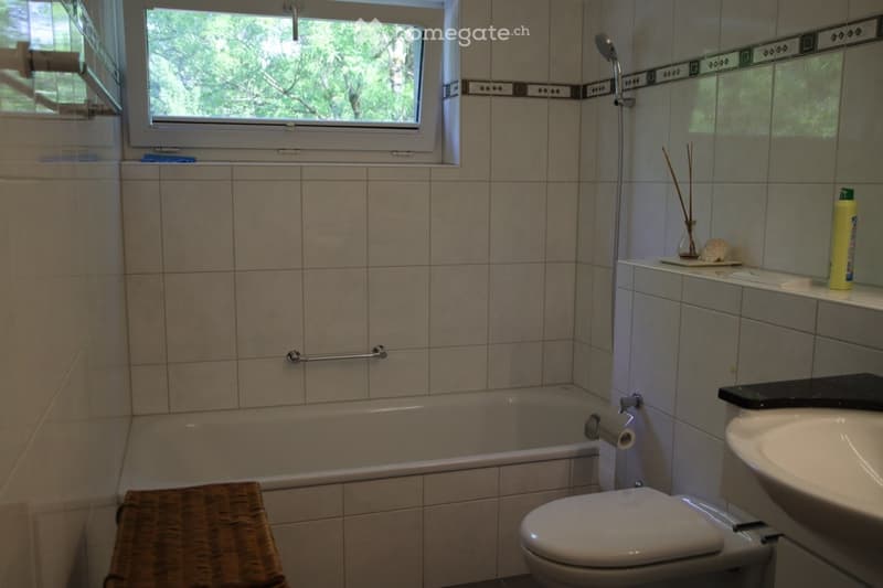Bathroom with tiled walls, bathtub, toilet, sink, wicker basket, and a window with a view of greenery.