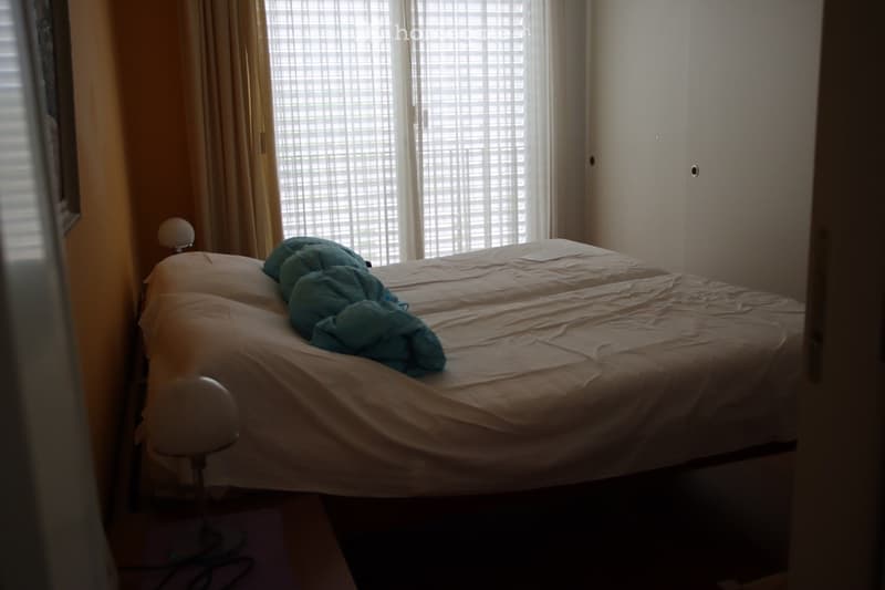 Bedroom with a single bed, white sheets, a blue blanket, and a bedside lamp near the window with blinds.