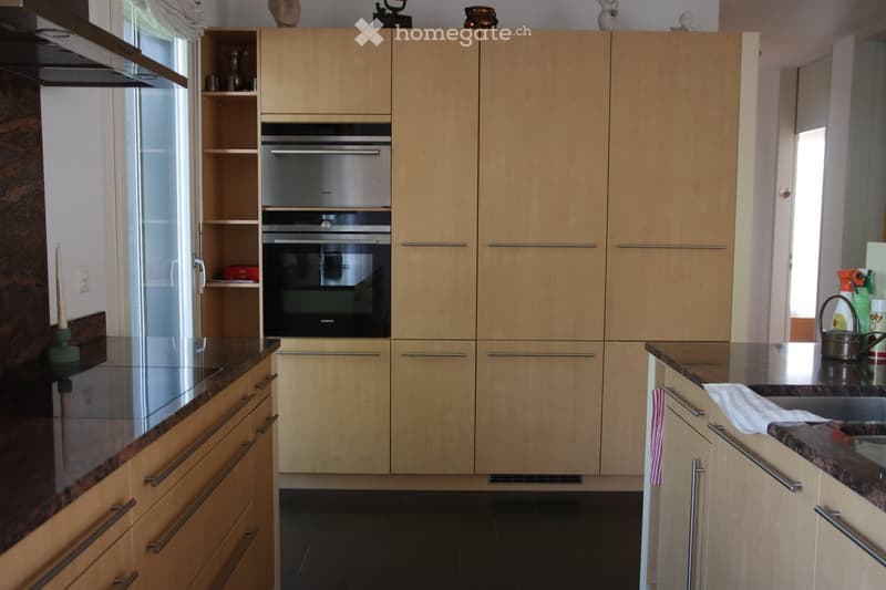 Modern kitchen with light wood cabinets, stainless steel oven, granite countertops, and a window with blinds.