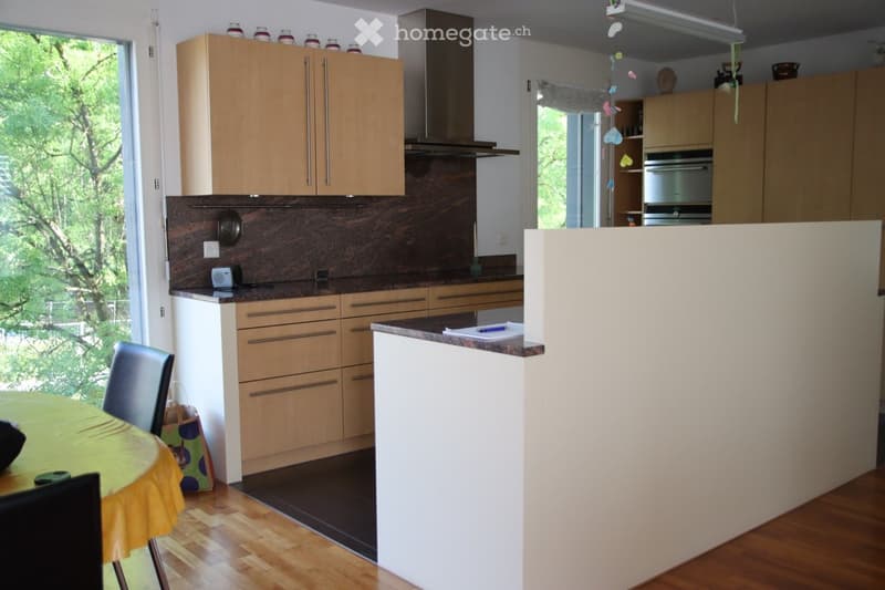 Modern kitchen with light wooden cabinets, stainless steel appliances, and a central island. The space includes a dining area with a round table and chairs.