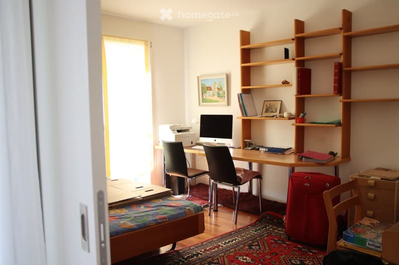 A furnished room with a bed, desk, chair, laptop, printer, bookshelf, suitcase, and picture frame. The room has wooden flooring and a colorful rug.