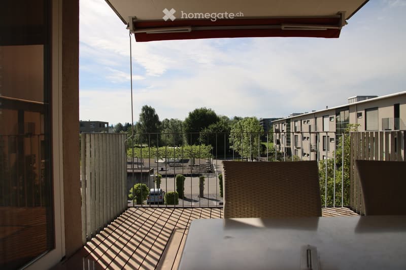 Balcony with wooden floor, metal railing, and outdoor furniture including a table and chair, overlooking a parking area with trees and buildings.