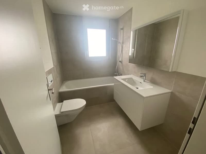 Modern bathroom with a white sink, bathtub, toilet, and a large mirror above the sink. The walls are tiled in neutral tones, and the floor is covered with beige tiles.