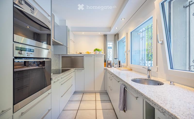 Modern kitchen with white cabinets, stainless steel appliances, granite countertops, and large windows. Features an oven, sink, blender, and a towel hanging on a rack.