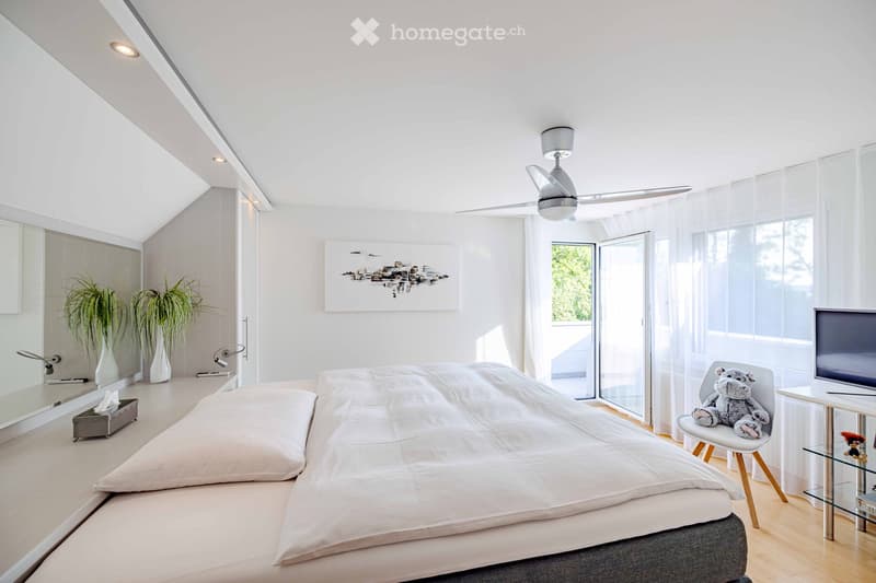 Modern bedroom with a large bed, white bedding, a chair with a stuffed animal, a small table with a TV, a ceiling fan, and decorative plants.