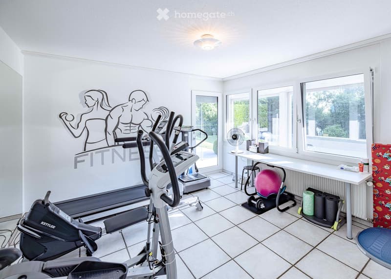 Well-lit fitness room with tiled floor, large windows, and various exercise equipment including a treadmill, elliptical machine, exercise bike, and a pink exercise ball. A small table with a fan and other items is also present.