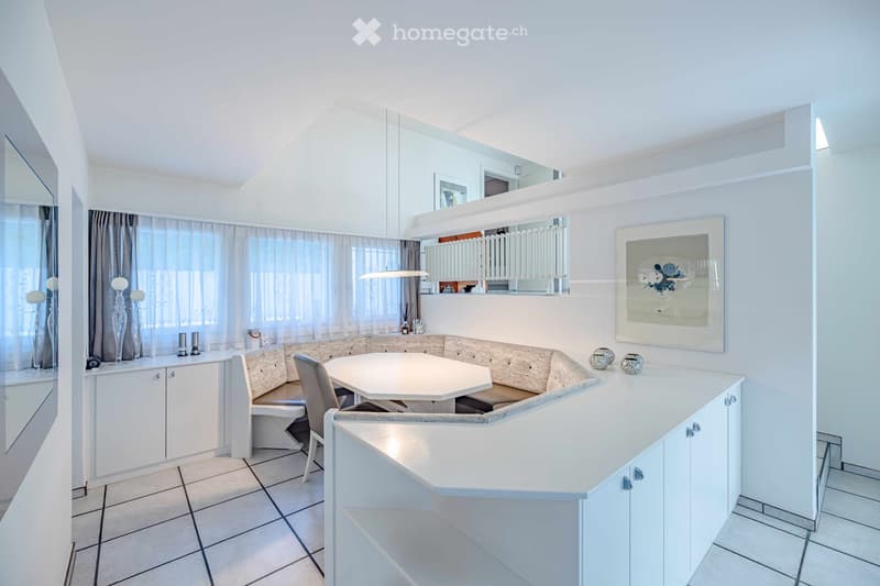 Bright, modern dining area with white cabinets, a round dining table, and a built-in bench seating area. The space features large windows with sheer curtains, decorative vases, and artwork on the walls.