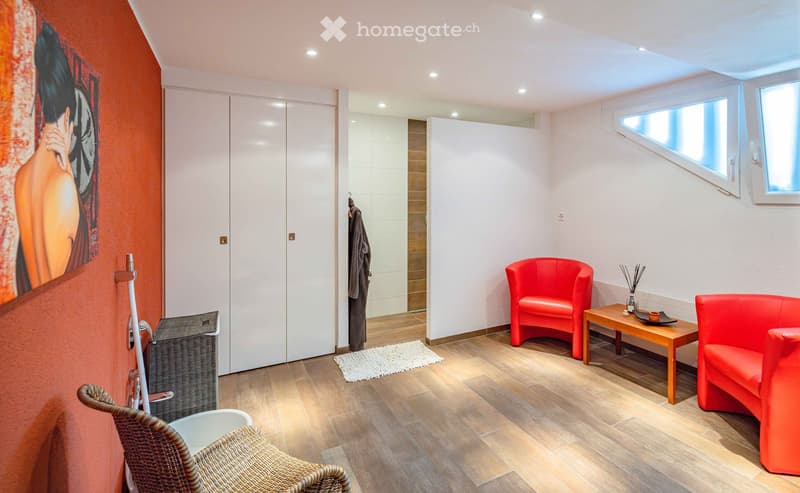 A modern, well-lit room with wooden flooring, red seating, a small table, and a large picture on the wall. The room features a closet, a small rug, and a wicker chair near the entrance.