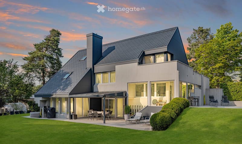 Modern two-story house with a dark grey roof and light grey walls, featuring large windows, a covered patio area, and a well-maintained lawn with outdoor furniture.
