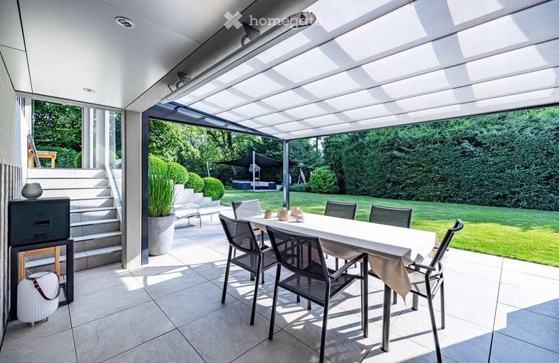 A modern, covered outdoor dining area with a white table, six chairs, and decorative vases, surrounded by greenery and a well-maintained lawn.