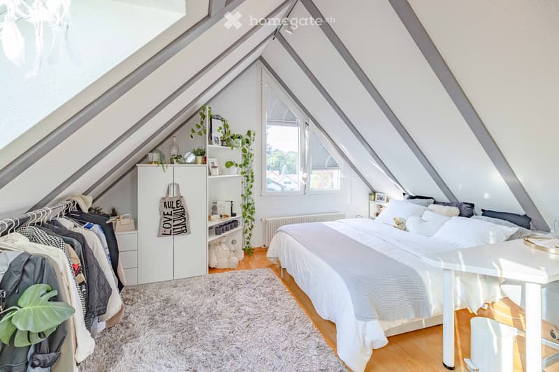 Cozy attic bedroom with slanted ceilings, large windows, a neatly made bed, a white wardrobe, a shelf with decorative items, and a small table with a chair.