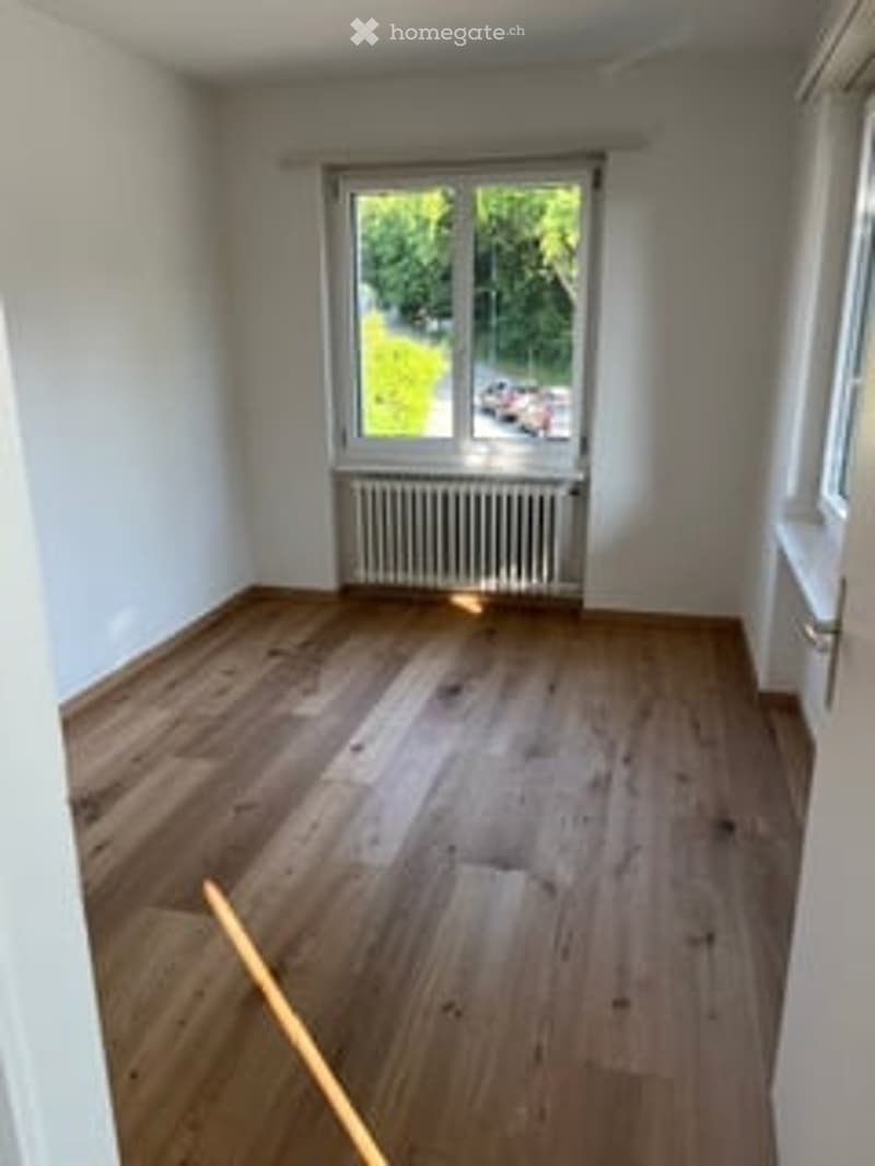 Completely empty room with wooden flooring, white walls, and a large window with a view of greenery outside.