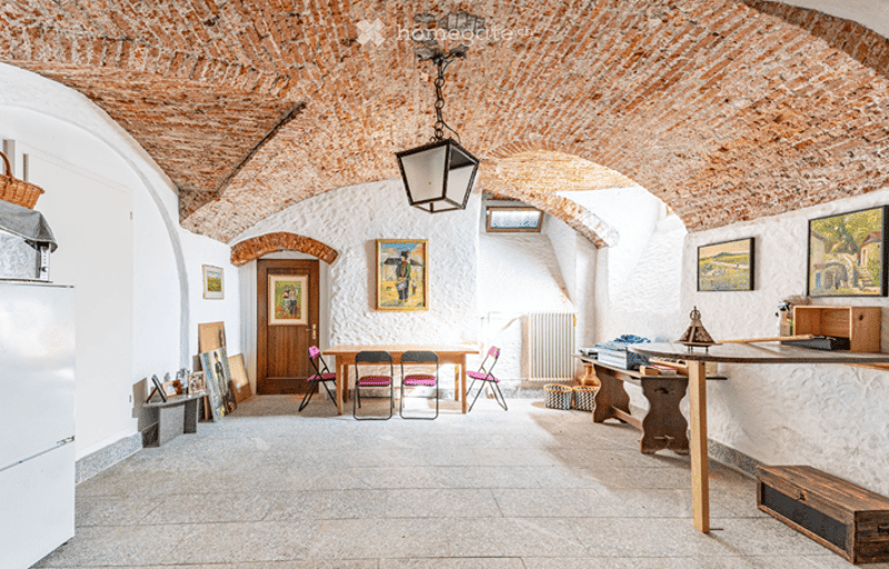 A spacious, high-ceilinged room with exposed brick arches and white walls. The room features a central wooden table surrounded by four pink chairs, a hanging lantern light fixture, several framed artworks on the walls, a wooden cabinet, and a small wooden