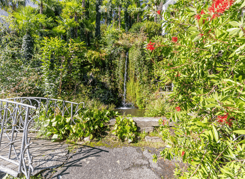 Lush garden with a small waterfall, greenery, and a metal railing leading to a pathway.