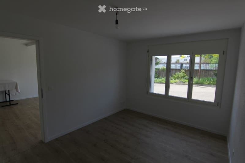 Empty room, white walls, hardwood floor, large window with outdoor view, small table in corner