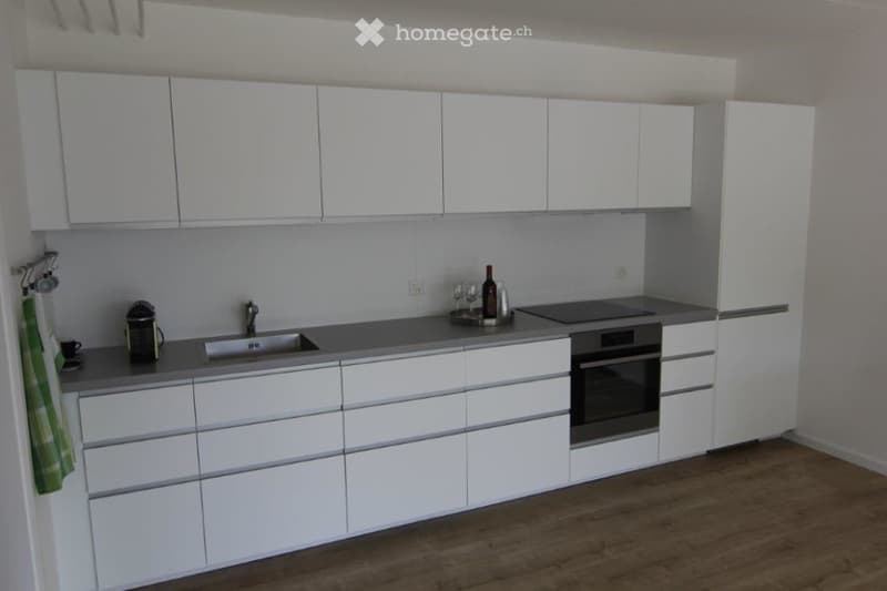 Modern kitchen with white cabinets, stainless steel appliances, and a grey countertop. Includes a sink, oven, refrigerator, and various small items like a coffee maker, wine glasses, and a bottle.