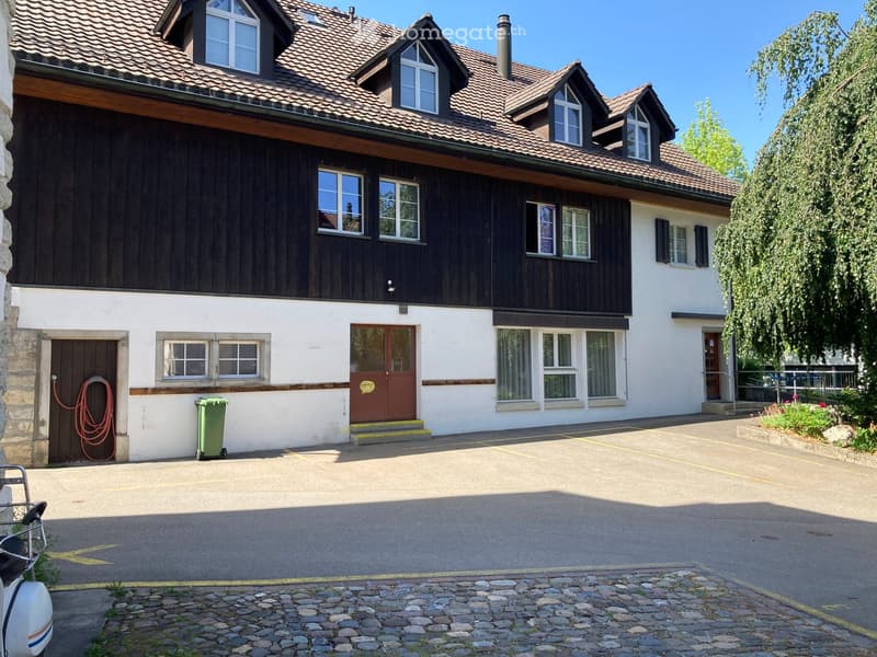 Two-story house with a tiled roof, wooden siding, and multiple windows. Features a central entrance with steps, a small garden area, and a paved driveway.