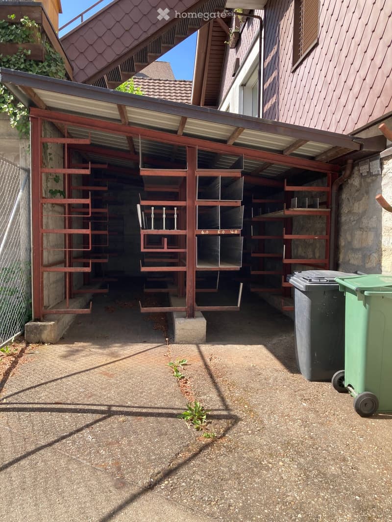 Outdoor area with a covered parking space, metal shelving units, and two trash bins.