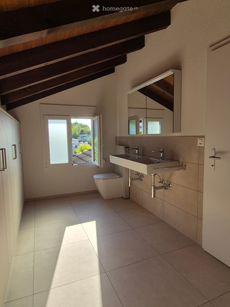 Modern bathroom with white tiled floor, white sink with dual faucets, toilet, large mirror, and exposed wooden beams on the ceiling. A window provides natural light.