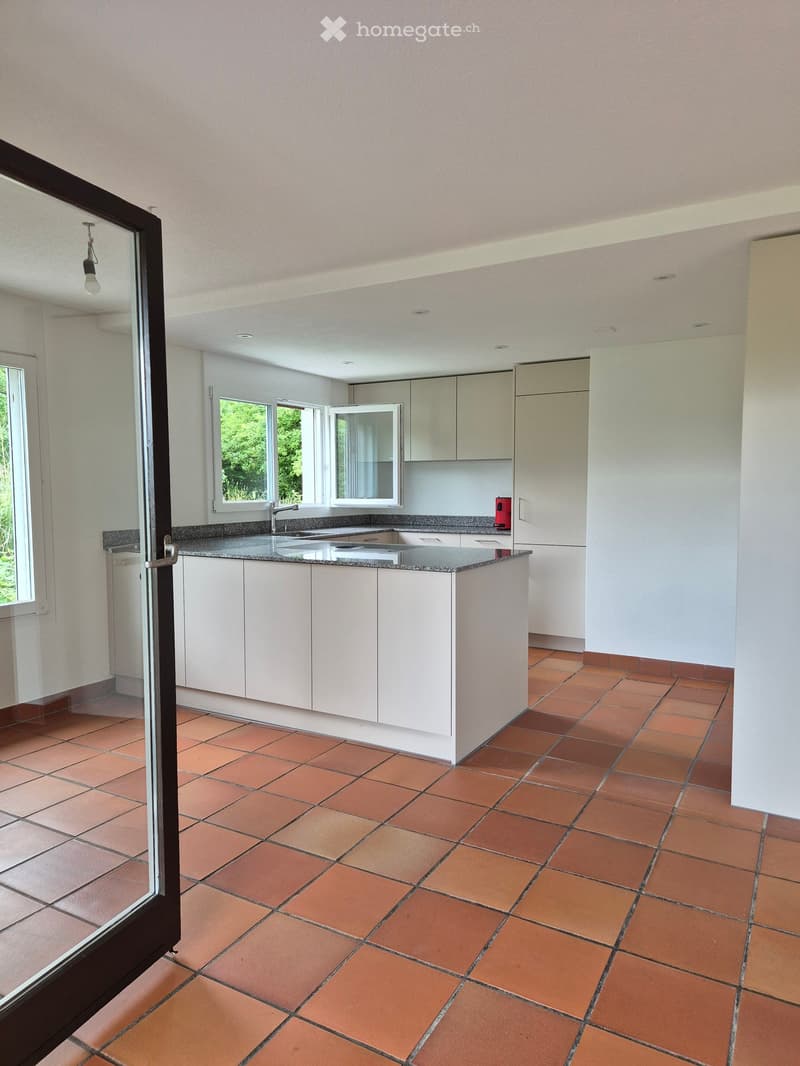 Modern kitchen with white cabinets, a central island, and terracotta tiled flooring. Features a sink with a faucet and large windows allowing natural light.