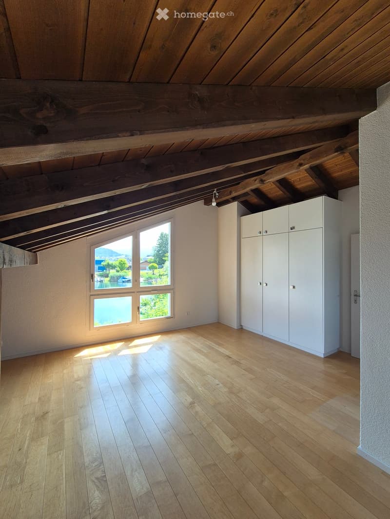 Empty room with wooden floor, white walls, exposed wooden ceiling beams, white built-in cabinets, and large window offering a scenic view of a lake and greenery.