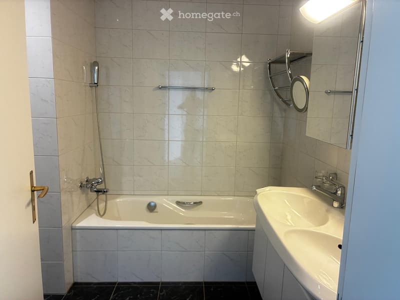 Modern bathroom with a white bathtub, a sink, and a mirror. The walls are tiled with marble-like tiles, and the floor is dark.