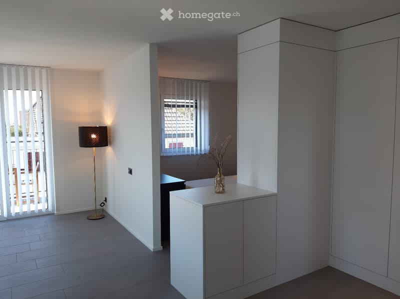 Minimalist room with white walls, tiled floor, a white cabinet with a vase, a lamp, and an open window with blinds. Adjacent room has built-in shelves and a window. Outside view of trees and buildings. A power outlet is on the wall.