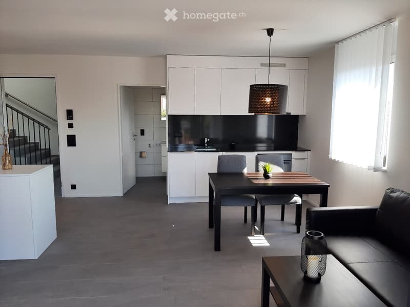Modern open floor plan with gray and white theme, dining area with table and chairs, kitchen with white cabinets and black countertops, living room with black couch and gray accent chair, chandelier and white blinds
