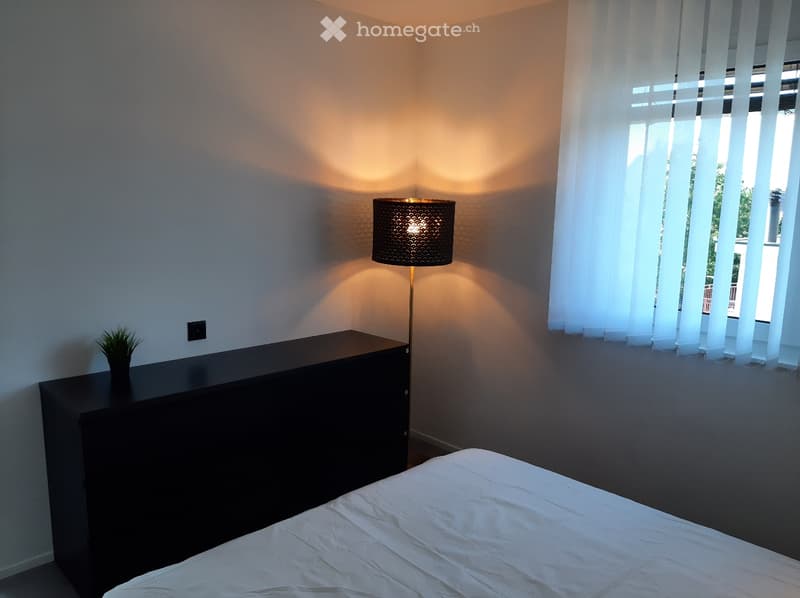 A room with a black cabinet and white bed, a lamp, white walls, and a window with blinds.