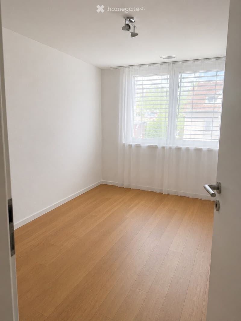 Empty room with light wooden flooring, white walls, large windows with blinds, and a door handle visible on the right side.