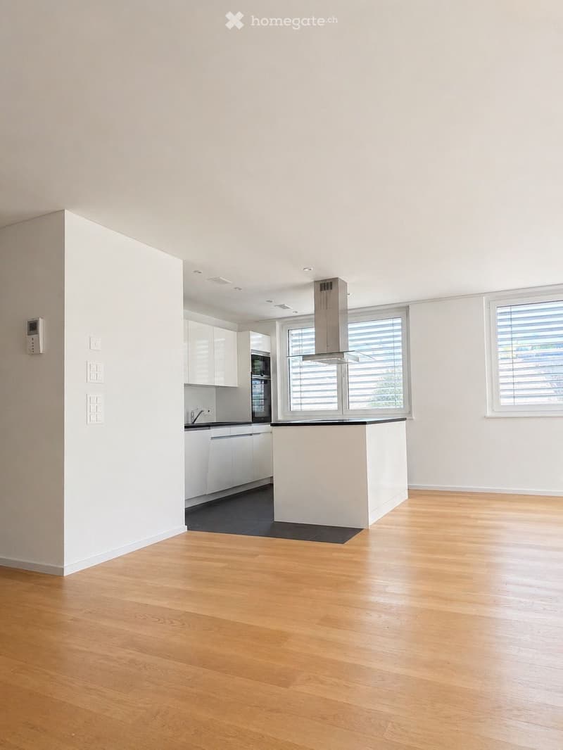 Open-plan kitchen and living area with light wooden flooring, white cabinetry, and large windows with blinds. The space is empty, with no furniture or appliances visible.